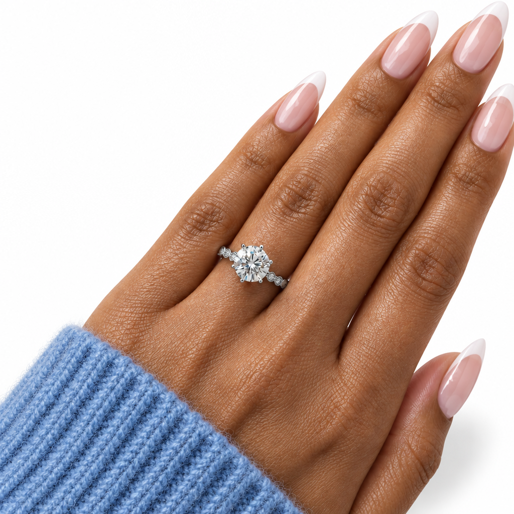 A close-up of a hand with a French manicure wearing a silver diamond engagement ring with a round-cut center stone and small side stones on a white background.