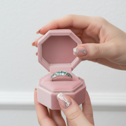 A silver ring with a green heart-shaped gemstone displayed in an open pink octagonal velvet ring box, held by two hands with decorated nails against a light gray background.