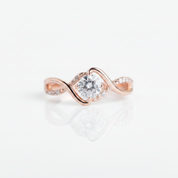 A rose gold engagement ring with a round-cut clear center stone and small accent stones along the band, shown on a clean white background with a soft reflection below.