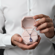 Person holding a pink jewelry box with a necklace featuring a purple teardrop pendant.
