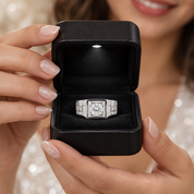 A silver-toned ring with a large round clear gemstone is displayed inside an open black jewelry box, held by a woman's hands against a softly blurred warm-toned background.