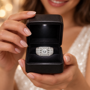 A silver ring with a large round center stone and surrounding pavé accents, displayed in an open black jewelry box held by a woman with manicured nails.