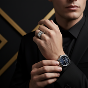 Man wearing a black suit with a watch and ring, against a dark background