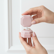 Hand holding a pink jewelry box with a ring inside on a light background