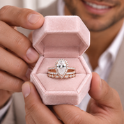 A close-up lifestyle shot of a man holding an open pink velvet ring box. Inside the box are a pear-cut diamond engagement ring with a halo of small diamonds and a matching diamond wedding band with rose gold-toned side stones.