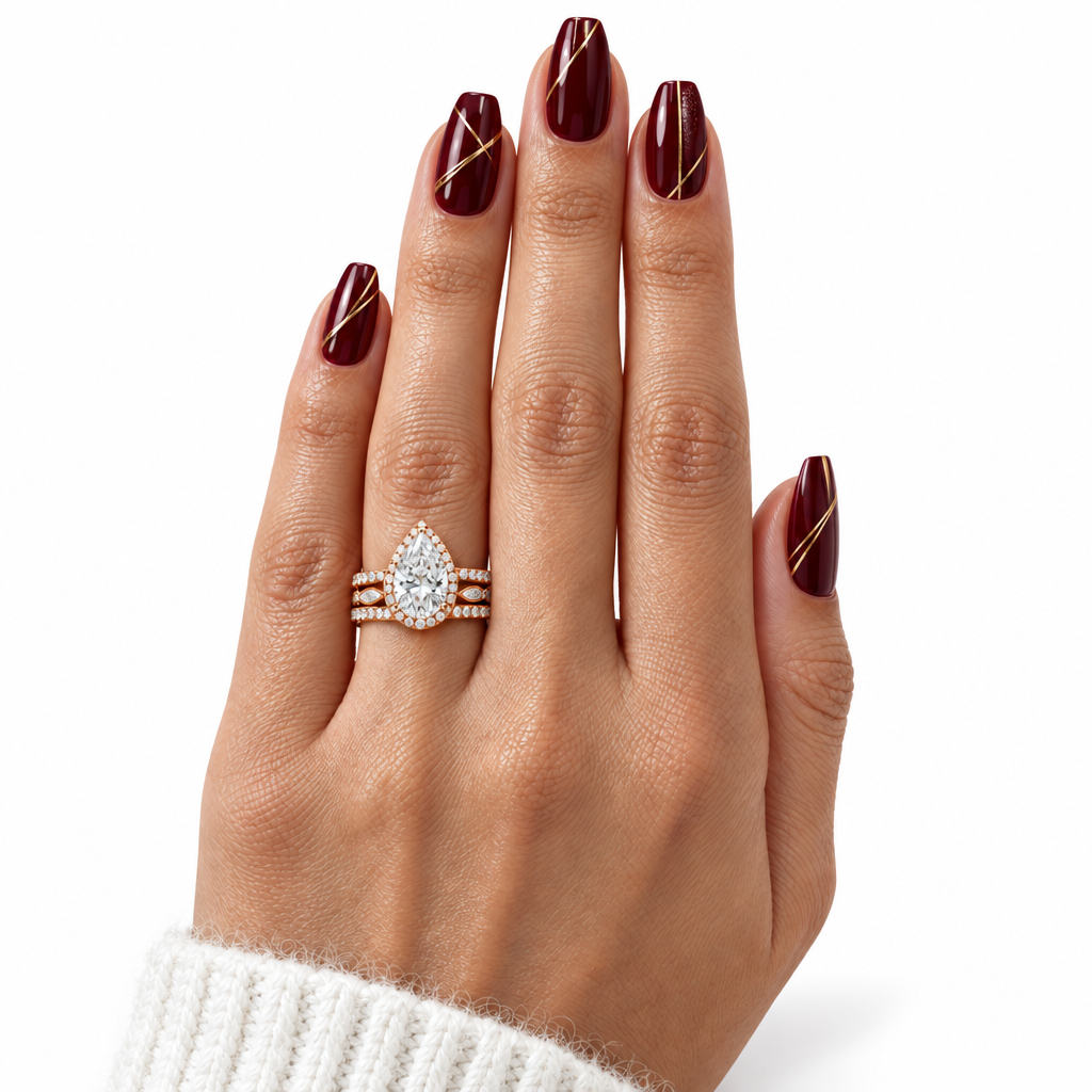 A close-up of a hand with dark burgundy nail polish wearing a three-stone ring set featuring a pear-cut clear center stone and small clear side stones in rose gold and white gold, against a clean white background.