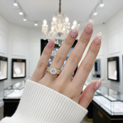 A close-up of a hand wearing a gold ring with a large clear center stone and smaller side stones, set against a softly blurred jewelry store interior with a chandelier and display cases.