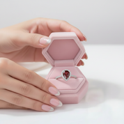 A silver ring with a teardrop-shaped red gemstone is displayed inside an open pink hexagonal jewelry box, held by two hands with light pink manicured nails and silver glitter tips.
