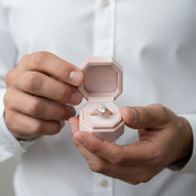 Person holding a pink jewelry box with an engagement ring inside, wearing a white shirt.