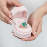 Pink jewelry box with a green gemstone ring held by hands on a light gray background