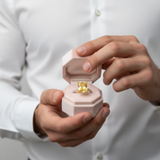 Person holding a pink jewelry box with a yellow diamond ring inside, wearing a white shirt.