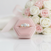 Gold ring with three diamonds in a pink hexagonal box, surrounded by pink and white roses.