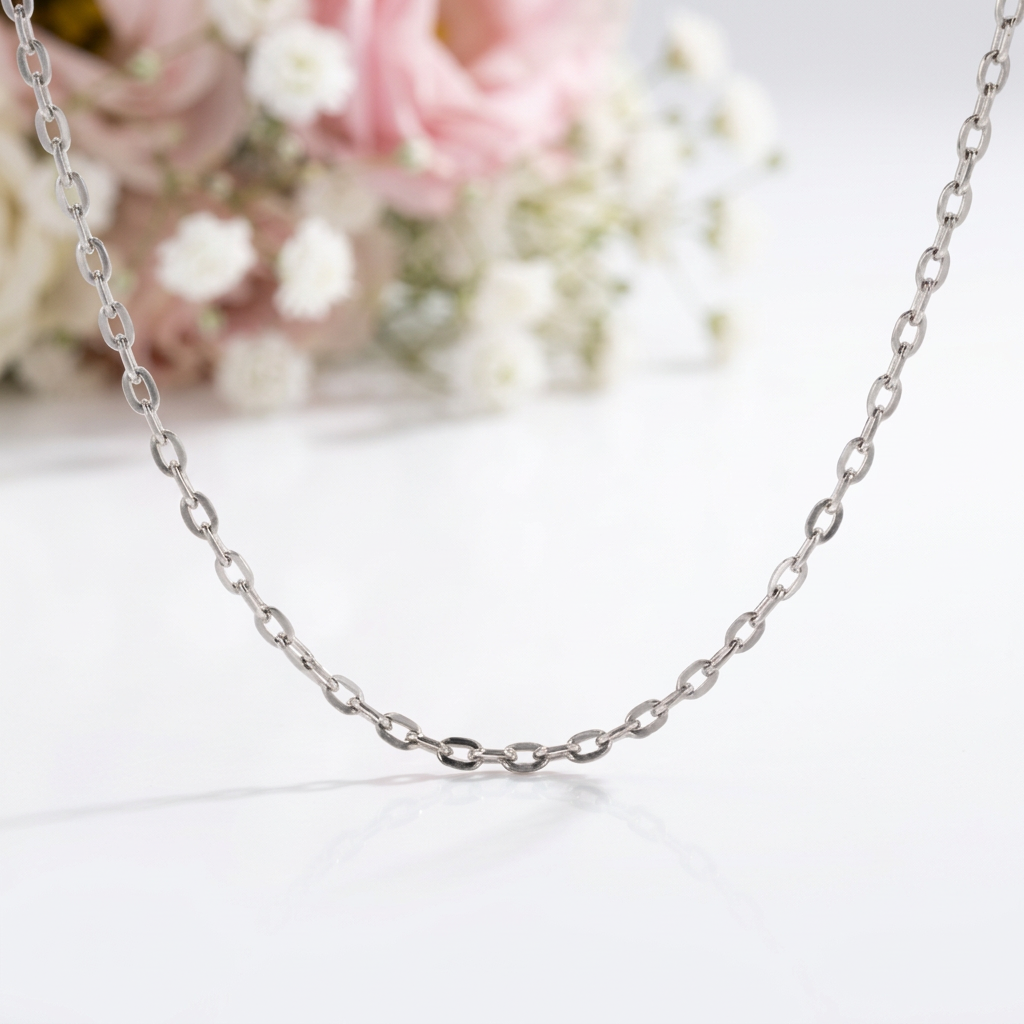 A silver-toned chain necklace with elongated oval links, displayed on a clean white surface with a soft-focus floral background of pink and white flowers.