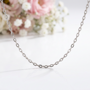 A silver-toned chain necklace with elongated oval links, displayed on a clean white surface with a soft-focus floral background of pink and white flowers.
