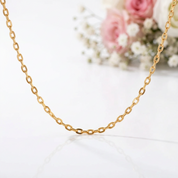 A delicate gold chain necklace draped in a U-shape against a clean white background, with soft pink and white flowers blurred in the background.