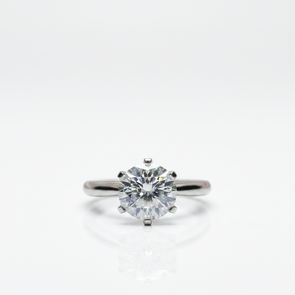 A classic solitaire engagement ring featuring a round brilliant-cut diamond set in a polished silver-colored metal band, shown on a clean white background with a soft reflection below.