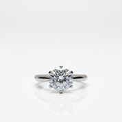 A classic solitaire engagement ring featuring a round brilliant-cut diamond set in a polished silver-colored metal band, shown on a clean white background with a soft reflection below.