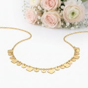 A gold chain necklace with multiple small geometric nameplate charms in different shapes, displayed on a white surface with soft pink roses in the background.