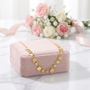 A delicate gold necklace with small geometric charms is draped over a pink velvet jewelry box on a white marble surface, with a blurred bouquet of pink and white roses in the background.