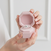 Hand holding a pink jewelry box with a silver ring inside on a light background