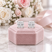 A pair of princess-cut diamond stud earrings displayed on a pink velvet jewelry box, with a soft-focus background of white and pink roses and a pink ribbon.