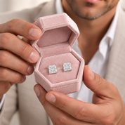 A close-up of a person holding an open pink jewelry box with two square-cut diamond stud earrings inside, against a softly blurred background of a man in a light suit.