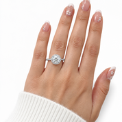 A close-up of a hand wearing a silver-toned halo ring with a round-cut center stone and smaller side stones, against a clean white background.