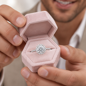 A close-up lifestyle shot of a man holding an open hexagonal pink velvet ring box, showcasing a round diamond engagement ring with a halo setting.