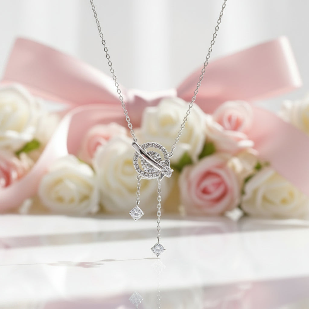 A silver-toned dangle necklace with a circular pavé pendant and two dangling clear stones, displayed against a soft pink ribbon and white roses background.