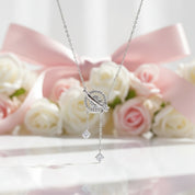A silver-toned dangle necklace with a circular pavé pendant and two dangling clear stones, displayed against a soft pink ribbon and white roses background.