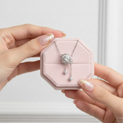 A silver-toned necklace with a planet ring pendant and dangling charms is displayed on a pink octagonal jewelry box held by two hands with manicured nails.