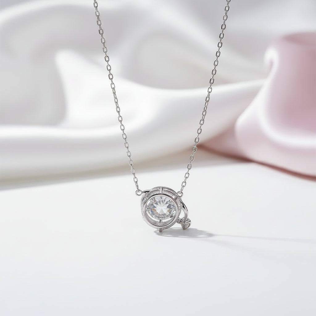 A silver necklace with a round bezel-set clear gemstone pendant, displayed on a clean white surface with soft pink and white satin fabric in the background.