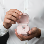 A close-up of a person in a white button-down shirt holding an open light pink jewelry box. Inside the box is a silver-toned necklace with a geometric pendant and matching drop earrings.