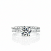 A silver-tone diamond ring set featuring a round-cut center stone and pavé-set bands, shown on a clean white background with a soft reflection below.