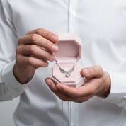 A person in a white dress shirt holds an open light pink jewelry box containing a silver angel-wing pendant necklace with a clear heart-shaped gemstone.