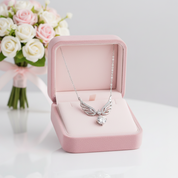 A silver angel-wing pendant necklace with a clear central gemstone is displayed inside an open pink jewelry box, with a bouquet of white and pink roses blurred in the background.