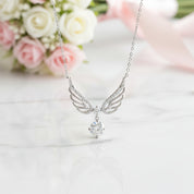 Silver angel wings pendant necklace with a clear round-cut gemstone hanging from the center, displayed on a white marble surface with soft pink and white roses in the background.