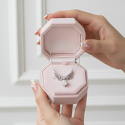 A silver angel-wing pendant necklace with a clear heart-shaped gemstone is displayed inside an open light pink octagonal jewelry box, held by two hands against a soft white background.