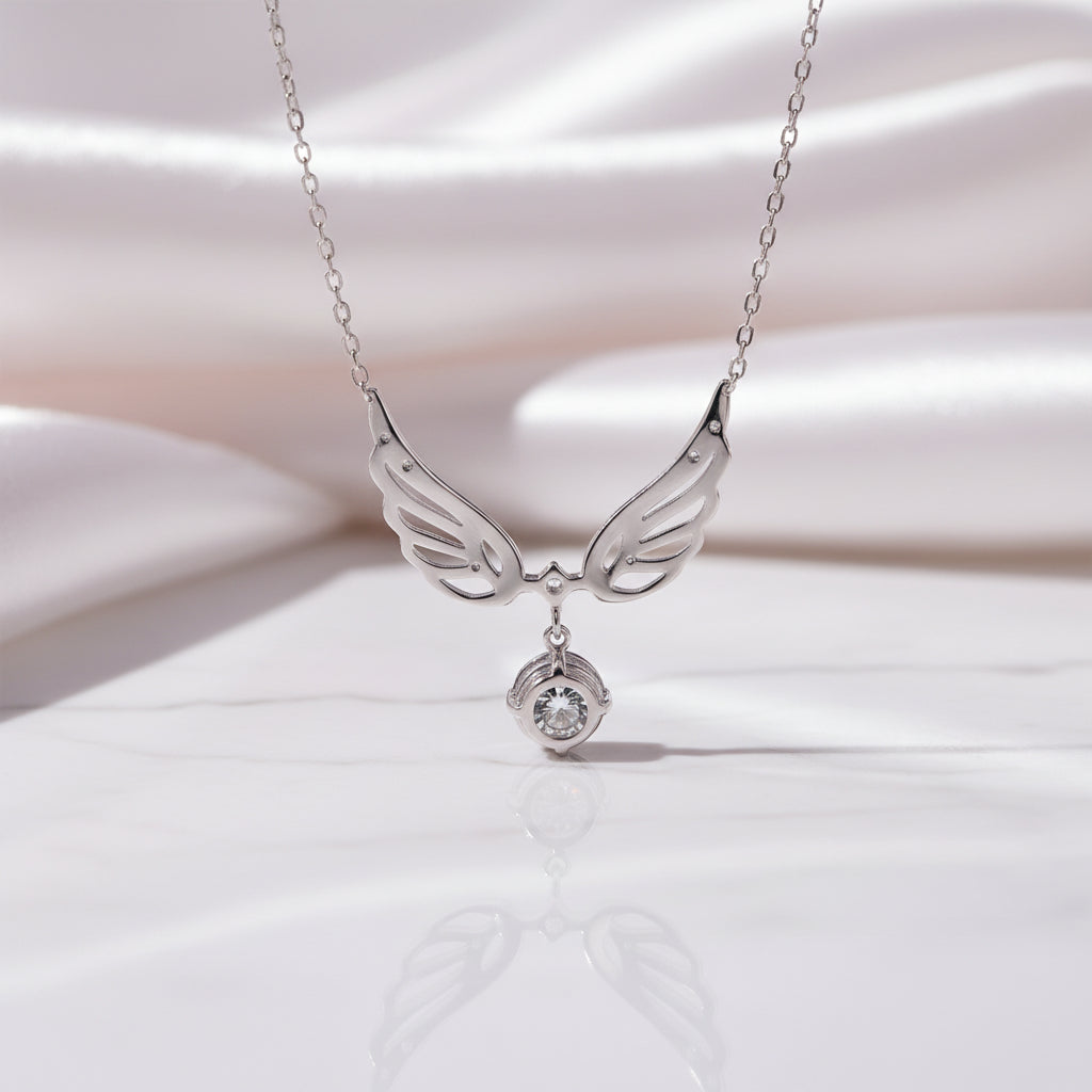 A silver angel wings pendant necklace with a small clear gemstone hanging below, displayed on a white marble surface with a soft white satin background.