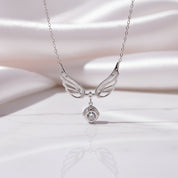 A silver angel wings pendant necklace with a small clear gemstone hanging below, displayed on a white marble surface with a soft white satin background.