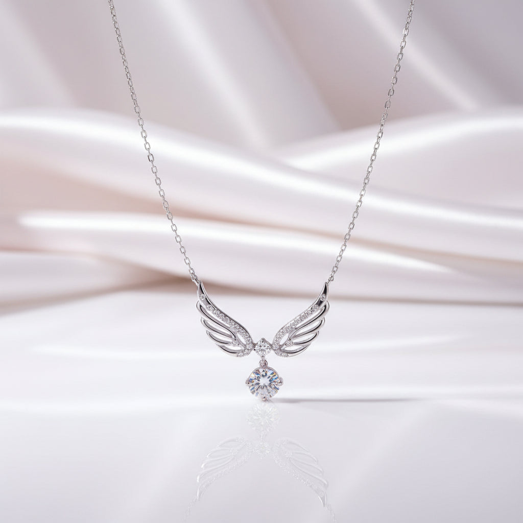 A silver angel wing pendant necklace with a sparkling clear gemstone, displayed on a soft, draped white satin fabric background.