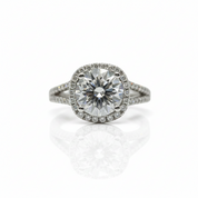 A round-cut halo ring featuring a large center stone surrounded by a halo of smaller stones, set on a split shank with pavé accents, photographed on a white background with a soft reflection below.