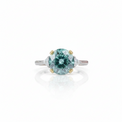 A center-stone ring featuring a round light blue-green gemstone in a four-prong setting, flanked by small clear side stones on a silver-toned band with gold accents, shown on a white background with a soft reflection.