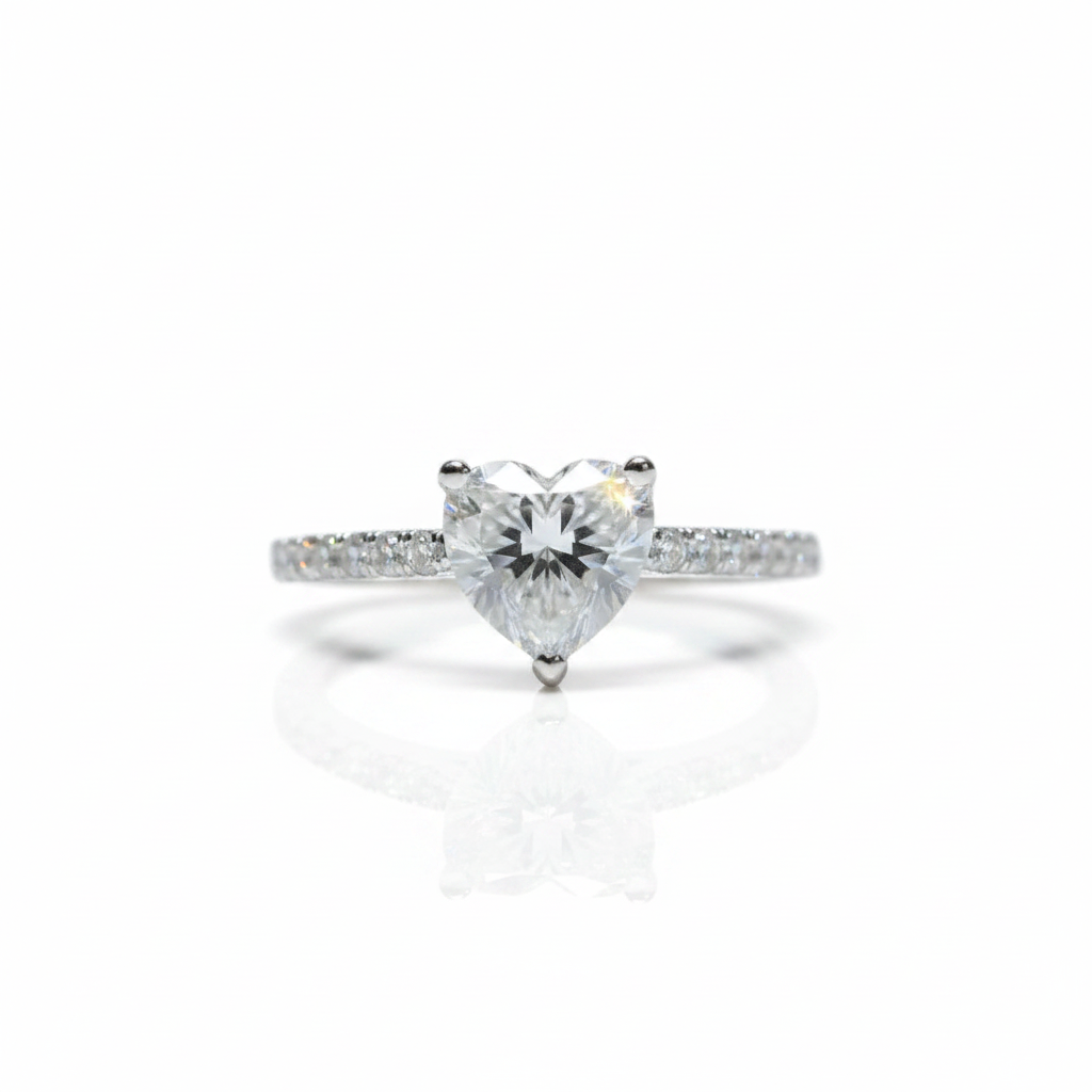 A heart-shaped diamond solitaire ring in a silver-colored setting, featuring a pavé band with small round diamonds, shown on a clean white background with a soft reflection.