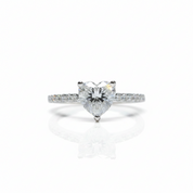 A heart-shaped diamond solitaire ring in a silver-colored setting, featuring a pavé band with small round diamonds, shown on a clean white background with a soft reflection.