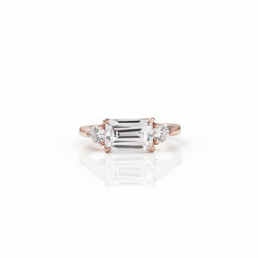 An emerald-cut diamond ring in a rose gold setting, flanked by two round diamonds, displayed on a clean white background with a soft reflection below.