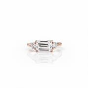 An emerald-cut diamond ring in a rose gold setting, flanked by two round diamonds, displayed on a clean white background with a soft reflection below.