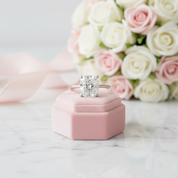 A radiant cut diamond engagement ring on a silver band, displayed on a pink velvet ring box with a pink rose bouquet in the background.