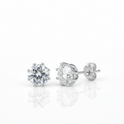 A pair of round-cut diamond stud earrings in a six-prong setting with polished silver-toned posts and backing, displayed on a reflective white surface.