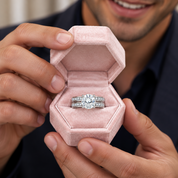 A close-up of a person holding an open pink velvet ring box with a silver-toned engagement ring featuring a round center stone and pavé band, with the person's hands and lower face visible.